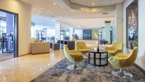 A modern lobby with a seating area featuring round tables and yellow chairs, a large window, and a reception desk.
