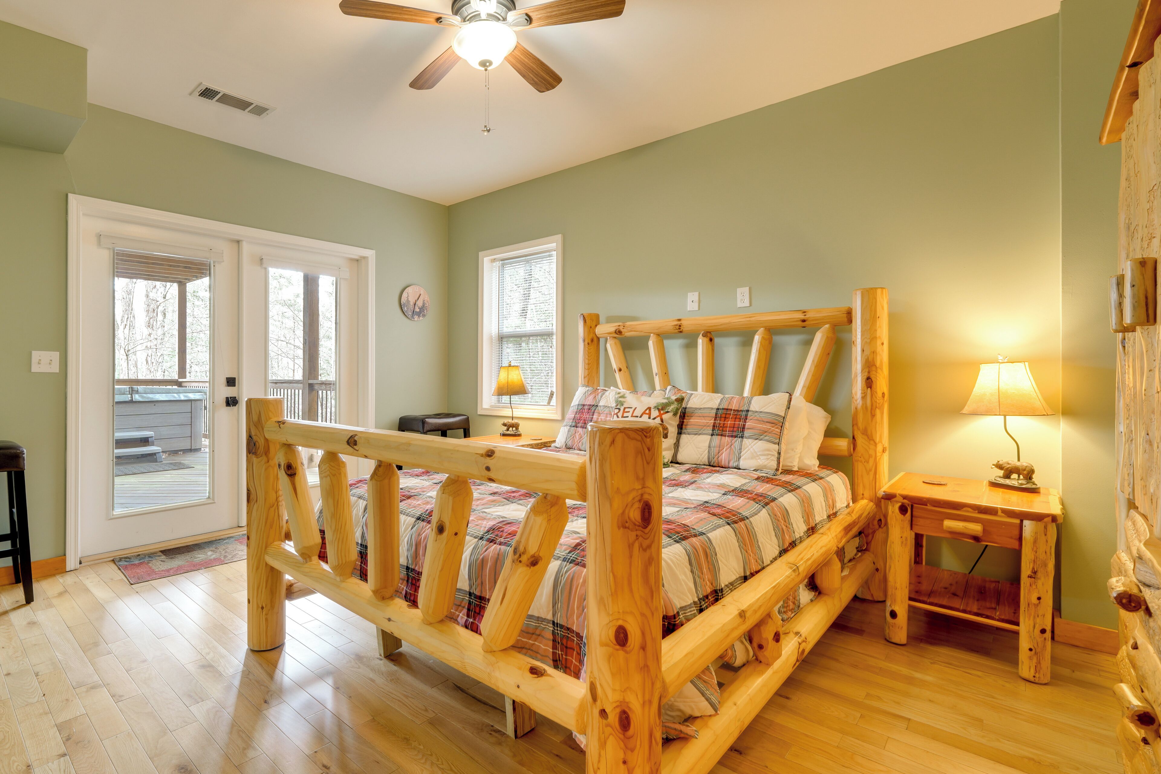 Una cama de madera con colcha escocesa, una mesita de noche con lámpara y un ventilador de techo en una habitación con paredes verdes y piso de madera.