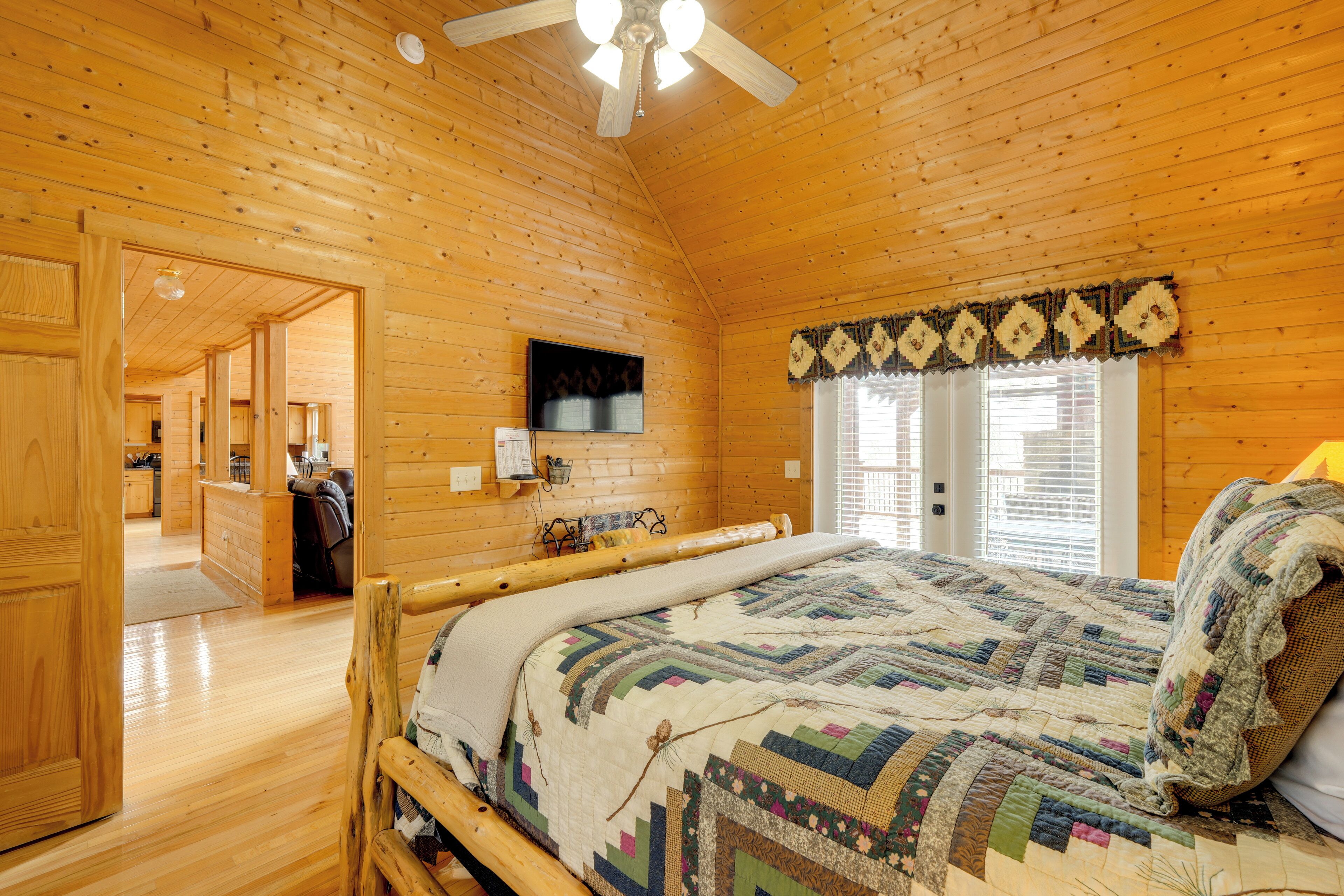Interior de una cabaña de madera con una cama, ventilador de techo, televisor y ventana con persianas.