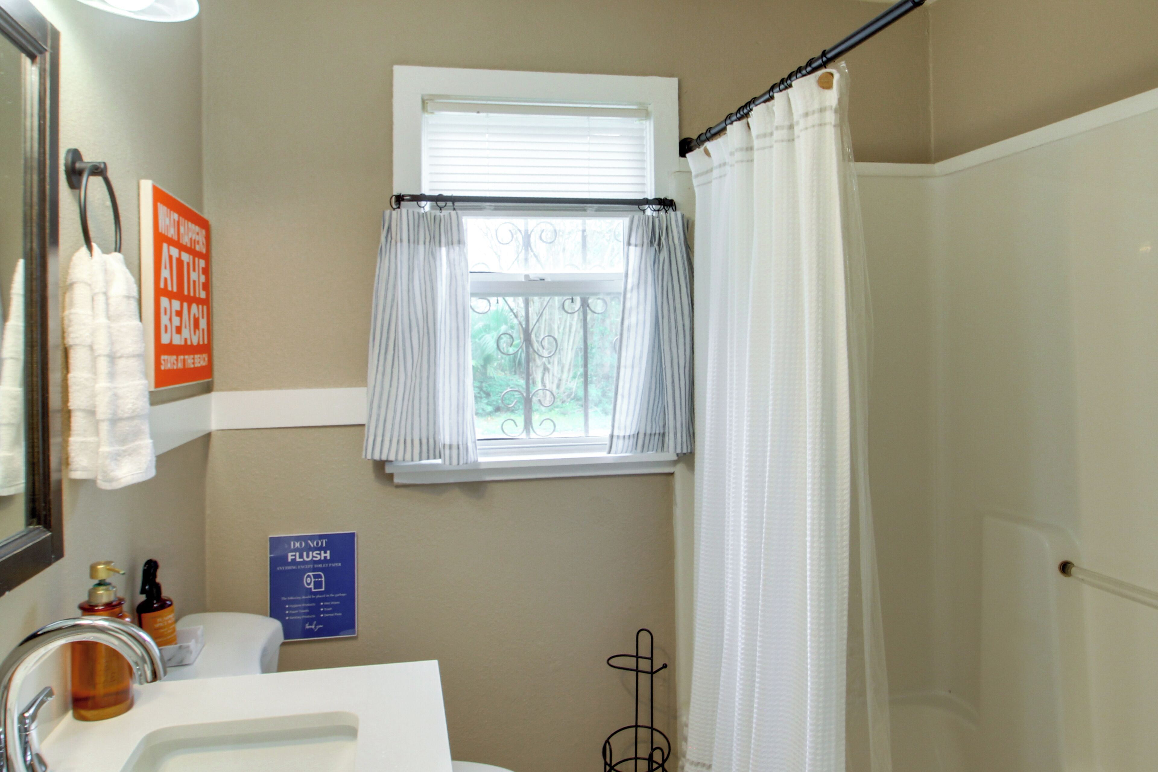 A bathroom with a sink, mirror, window with curtains, and a sign on the wall.