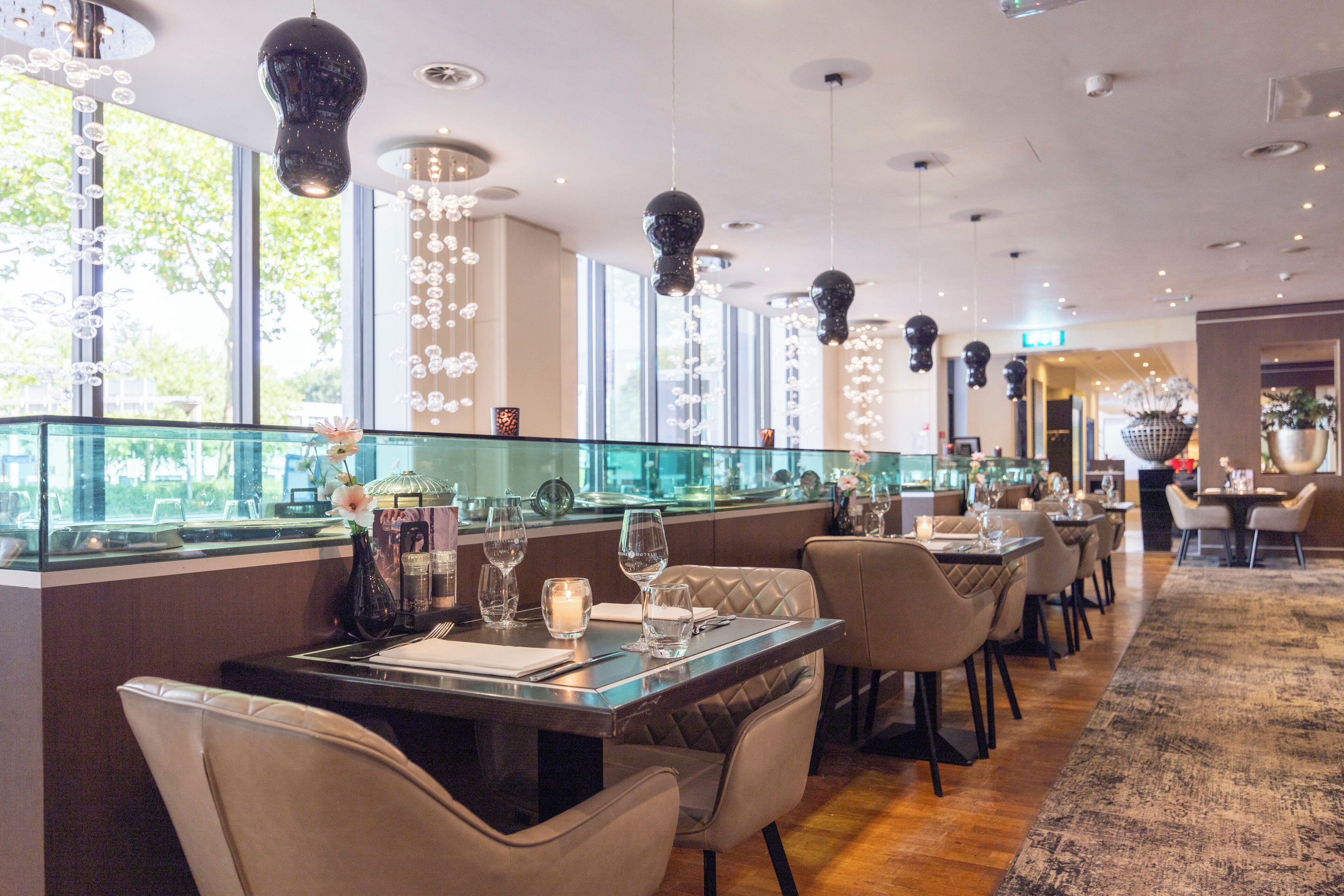 Un restaurante con divisiones de vidrio, mesas con sillas, iluminación colgante y grandes ventanales.