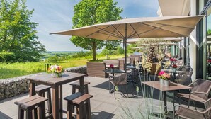 Un patio al aire libre con mesas y sillas, un paraguas grande y vistas al paisaje.