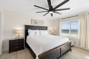 Une chambre à coucher comprenant un lit, une table de chevet, un ventilateur de plafond, une fenêtre avec des rideaux et une vue sur l’océan.