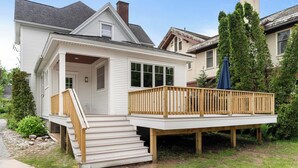A two-story house with a front porch, a wooden deck, and a staircase leading to the entrance. - Stylishly Renovated Retreat Just Steps from Dining & Entertainment (Petoskey)