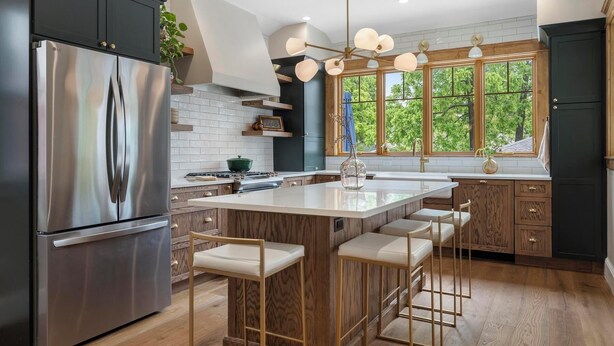 A modern kitchen with a stainless steel refrigerator, wooden cabinets, a central island with bar stools, and large windows offering a view of greenery. - Stylishly Renovated Retreat Just Steps from Dining & Entertainment (Petoskey)