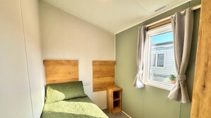Une petite chambre avec un lit simple, une couvre-lit verte, une tête de lit en bois, une petite table de chevet en bois et une fenêtre avec des rideaux.