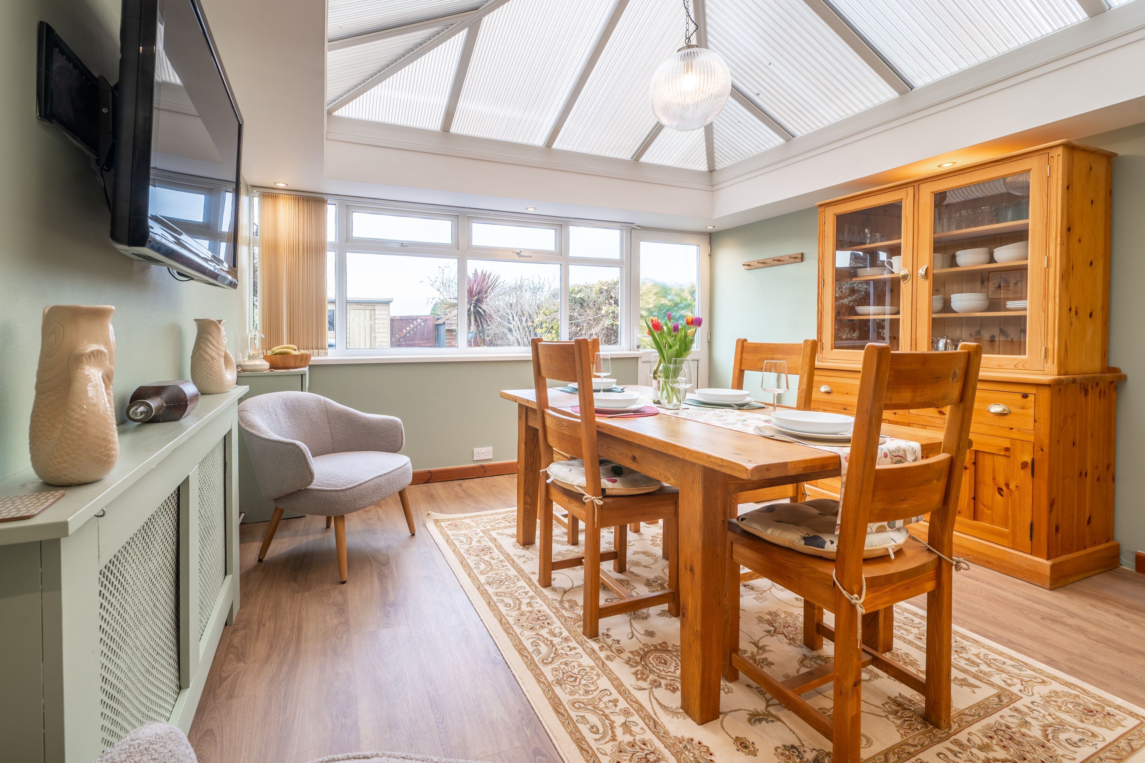 A dining area with a wooden table, chairs, a television mounted on the wall, and a large window with a view of the outside.