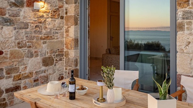 A stone exterior wall, a wooden dining table with chairs, a bottle, and a bowl of fruit. - Villa Alexia - Private Pool Panoramic Sea Views (Samos)