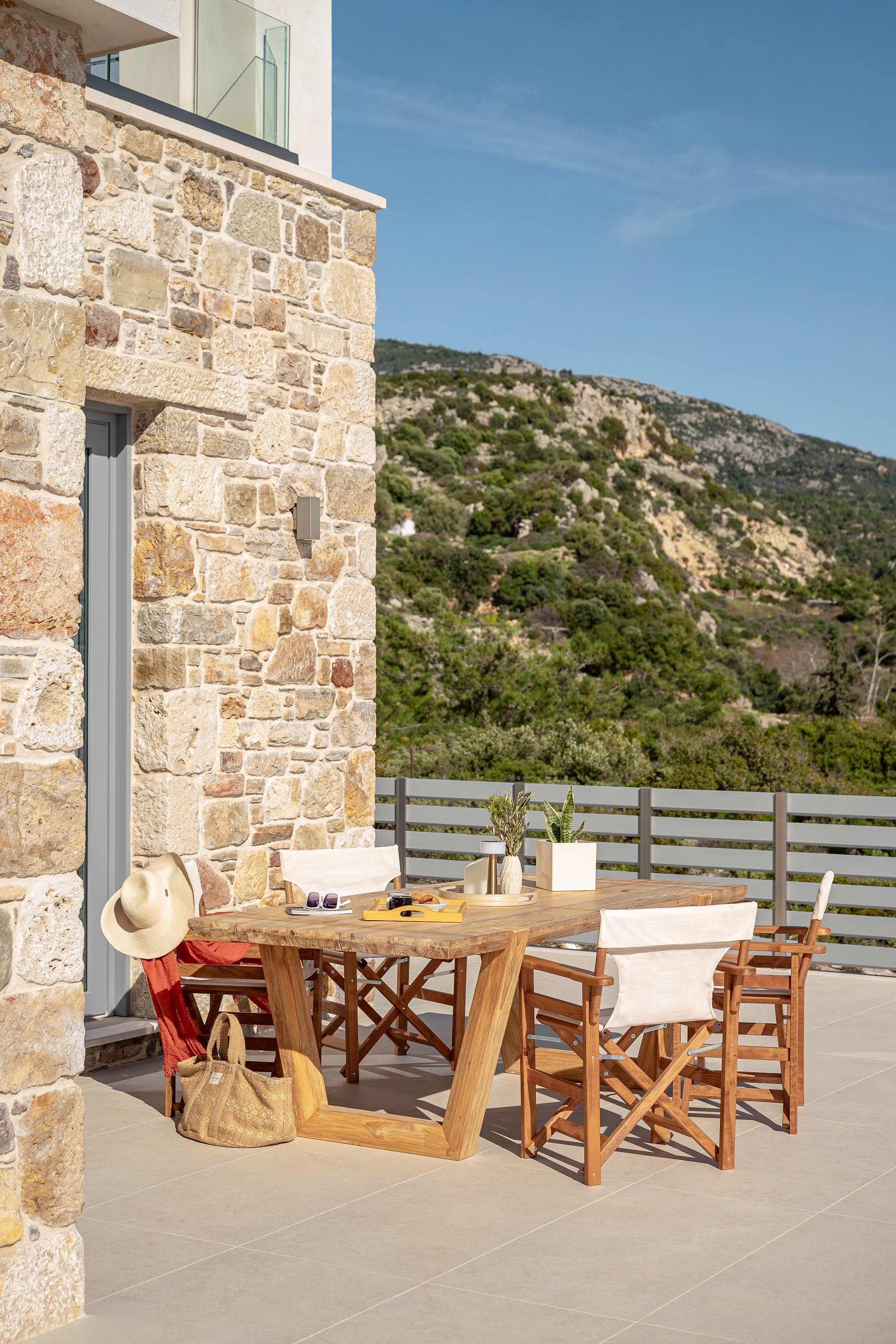 A stone wall, wooden table, and chairs on an outdoor terrace with a view of a mountainous landscape.