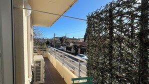 A balcony with a green table, a white air conditioning unit, a beige awning, and a view of a street with houses and a palm tree. - Le Chic Azuréen (Villeneuve-Loubet)