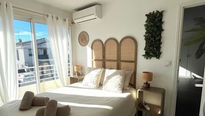 A bedroom with a large bed, a bedside table with a lamp, a wall-mounted air conditioner, and a view of the outside. - Le Chic Azuréen (Villeneuve-Loubet)