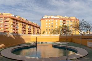 A swimming pool surrounded by a yellow building with balconies, a clear sky, and a few trees. - Vacation Apartment in Roquetas de Mar (Roquetas de Mar)