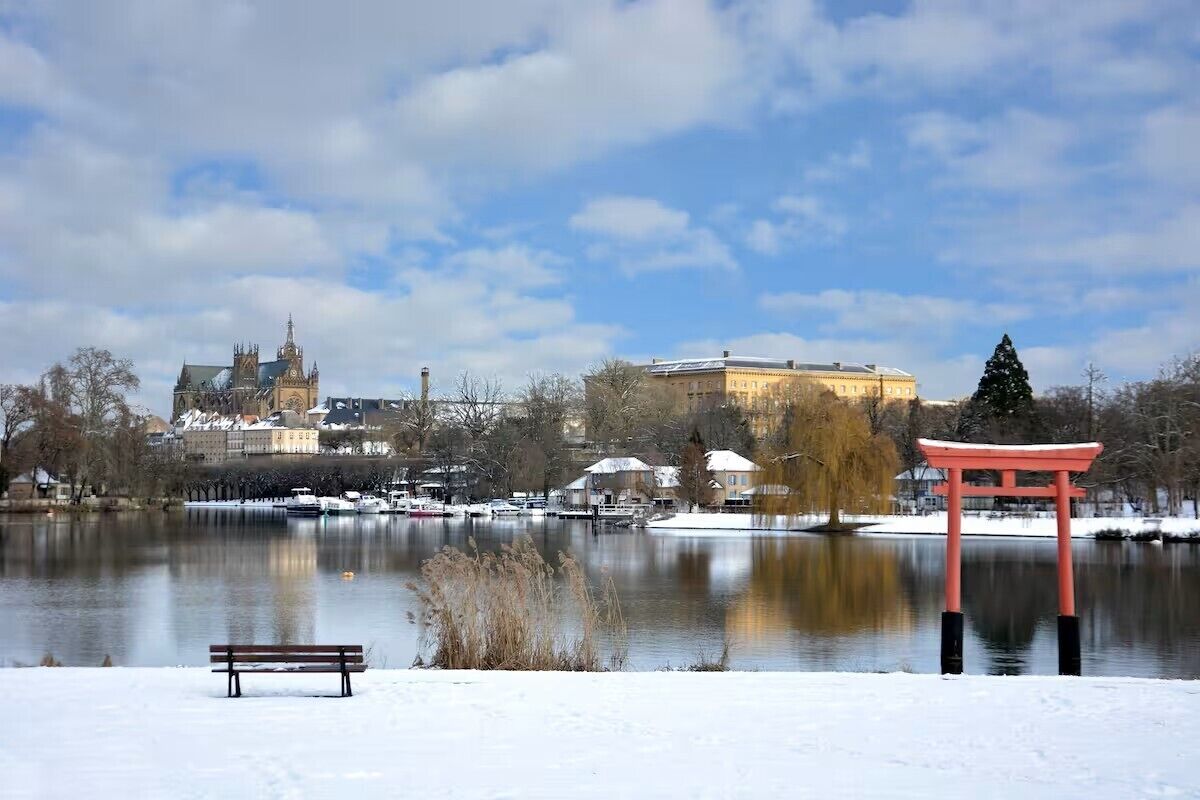 Fredfyldt søudsigt med en traditionel japansk torii-port, en bænk og historiske bygninger i baggrunden.