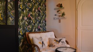 Ein Zimmer mit tropischer Wandgestaltung, ein gepolsterter Sessel mit Zierkissen, ein kleiner runder Tisch und eine Tür.
