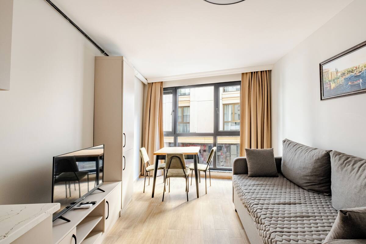 Una sala moderna con sofá, una mesa de comedor con sillas, un televisor sobre un mueble bajo y un ventanal con cortinas.
