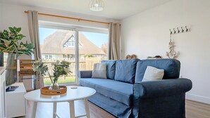 Ein Wohnzimmer mit einem blauen Sofa, einem weißen Tisch, einer Pflanze und Blick auf ein Haus durch ein großes Fenster.