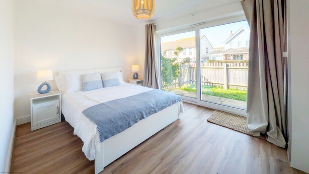 Ein Schlafzimmer mit einem großen Bett, zwei Nachttischen, einer Pendelleuchte und Blick auf einen Garten und Häuser durch eine Glastür.