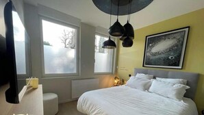 A bedroom with a bed, bedside table, two candles, a round stool, a window, and a wall-mounted artwork.