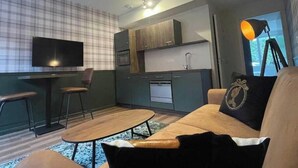 A modern living room with a sofa, coffee table, and a TV. There is a kitchen area with cabinets and a sink.