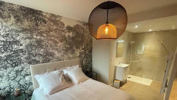 A modern bedroom with a large bed, a wall mural, a hanging light fixture, a glass shower enclosure, and a small bedside table.
