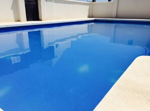 Ein klarblauer Swimmingpool mit weißem Rand, einer Betonplattform und einer weißen Wand mit einem schwarzen Lüftungsgitter.