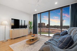 A modern living room with a large flat-screen TV, a wooden coffee table, a sofa with decorative pillows, a floor lamp, and a view of a stadium through large windows. - Massive Stadium View | 2BR | Free Parking|CBD (Kangaroo Point)