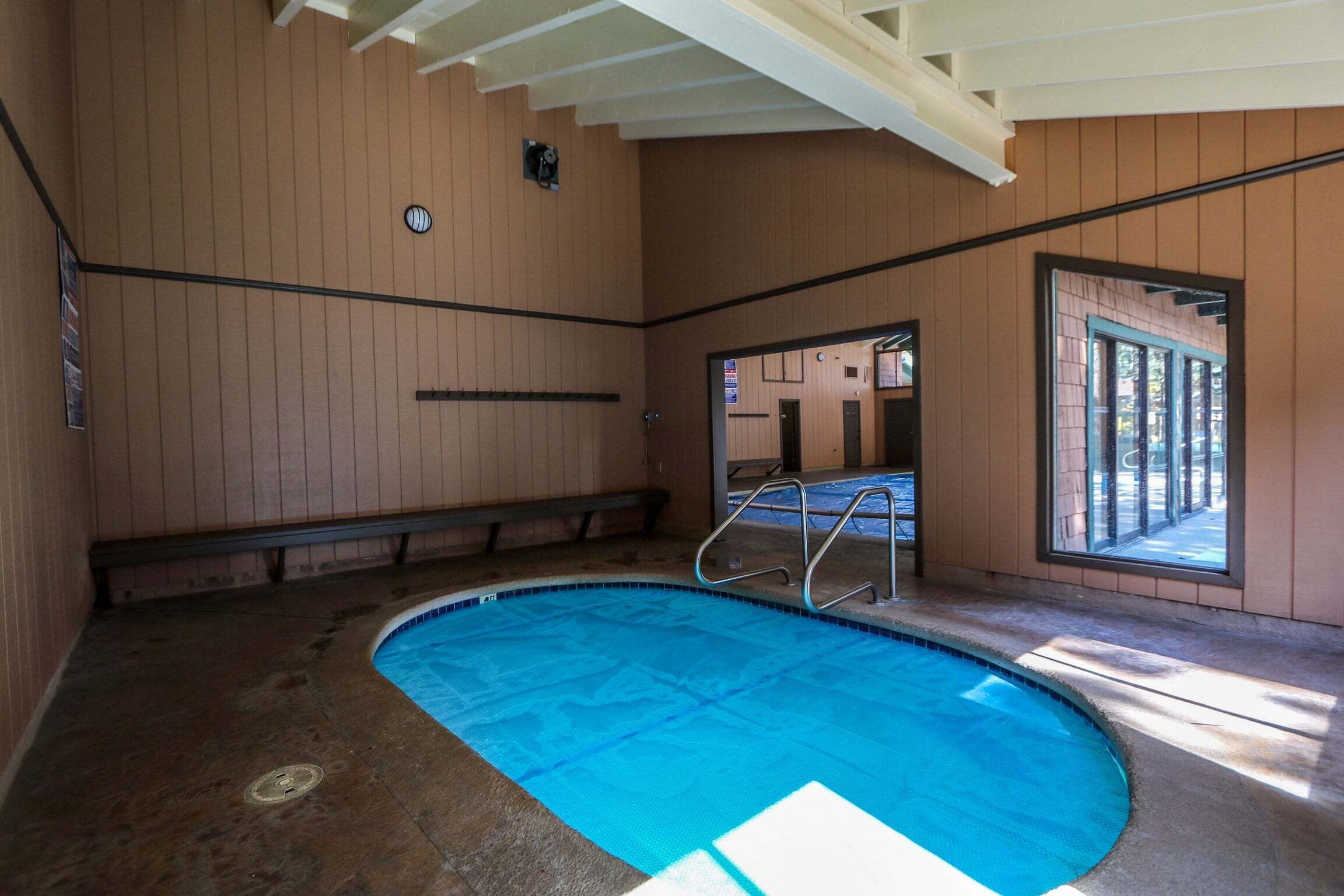 Piscina interior com paredes de madeira, escada de metal e vista pela janela.