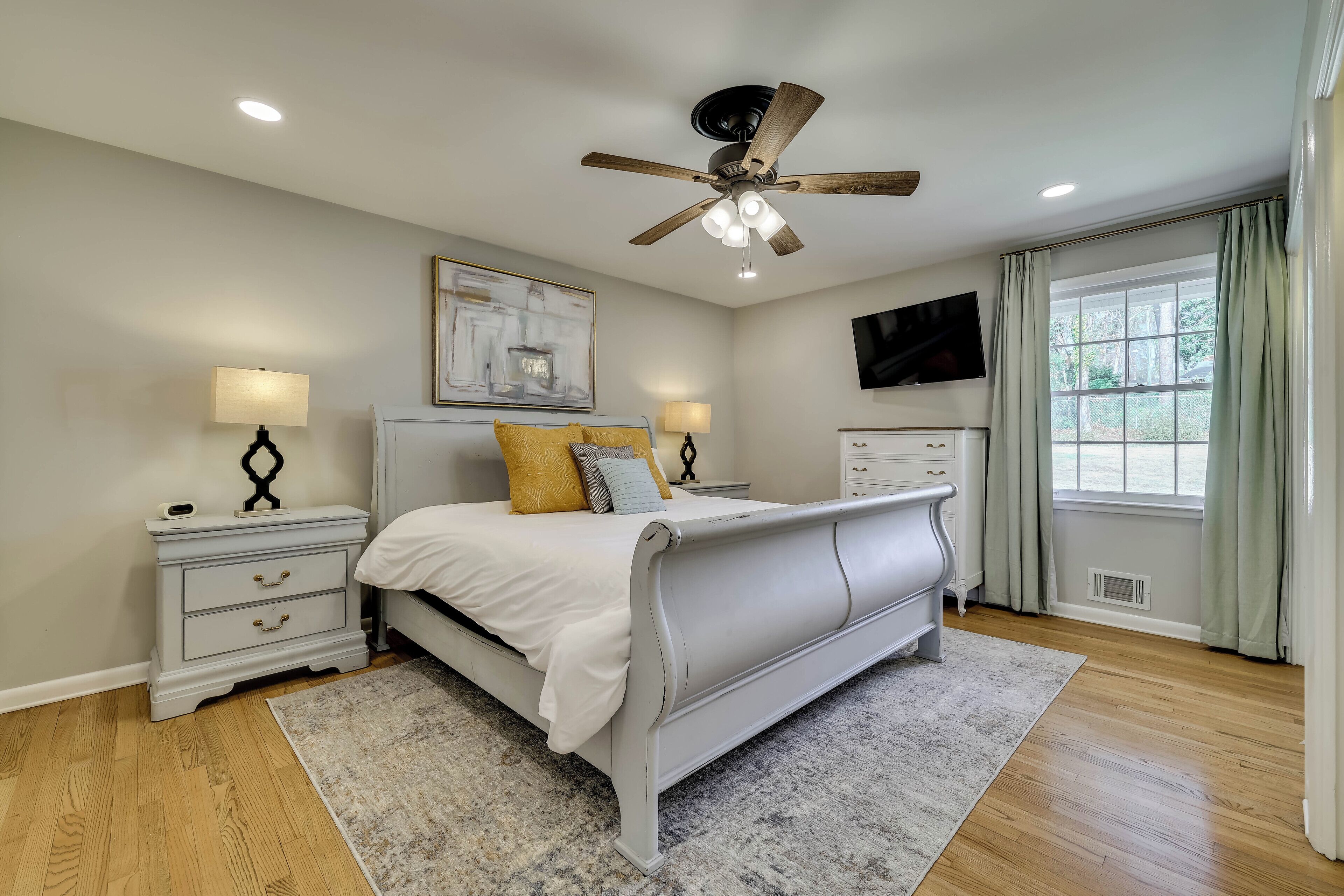 A bedroom with a bed, nightstands, a ceiling fan, a TV, and a window with curtains.