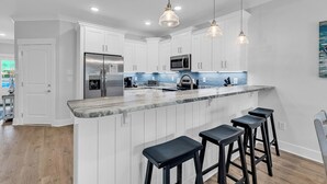 Eine moderne Küche mit weißen Schränken, einem Edelstahl-Kühlschrank, einer Mikrowelle und einer Marmorarbeitsplatte. Drei schwarze Barhocker und Pendelleuchten befinden sich über der Arbeitsfläche.