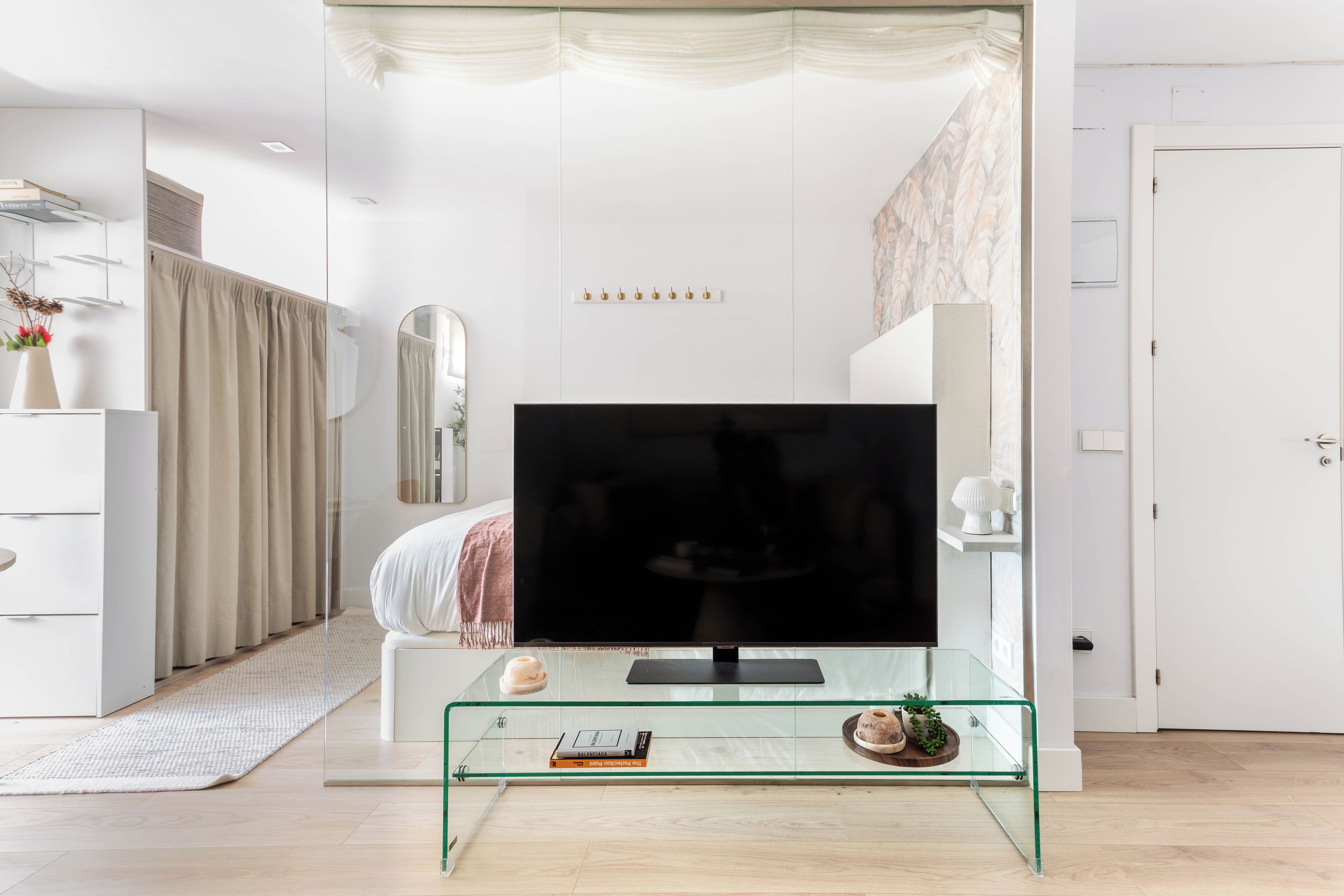 Un dormitorio moderno con un mueble de televisión de vidrio, una cama con manta blanca y rosa, una mesita de noche con un libro y un espejo grande.