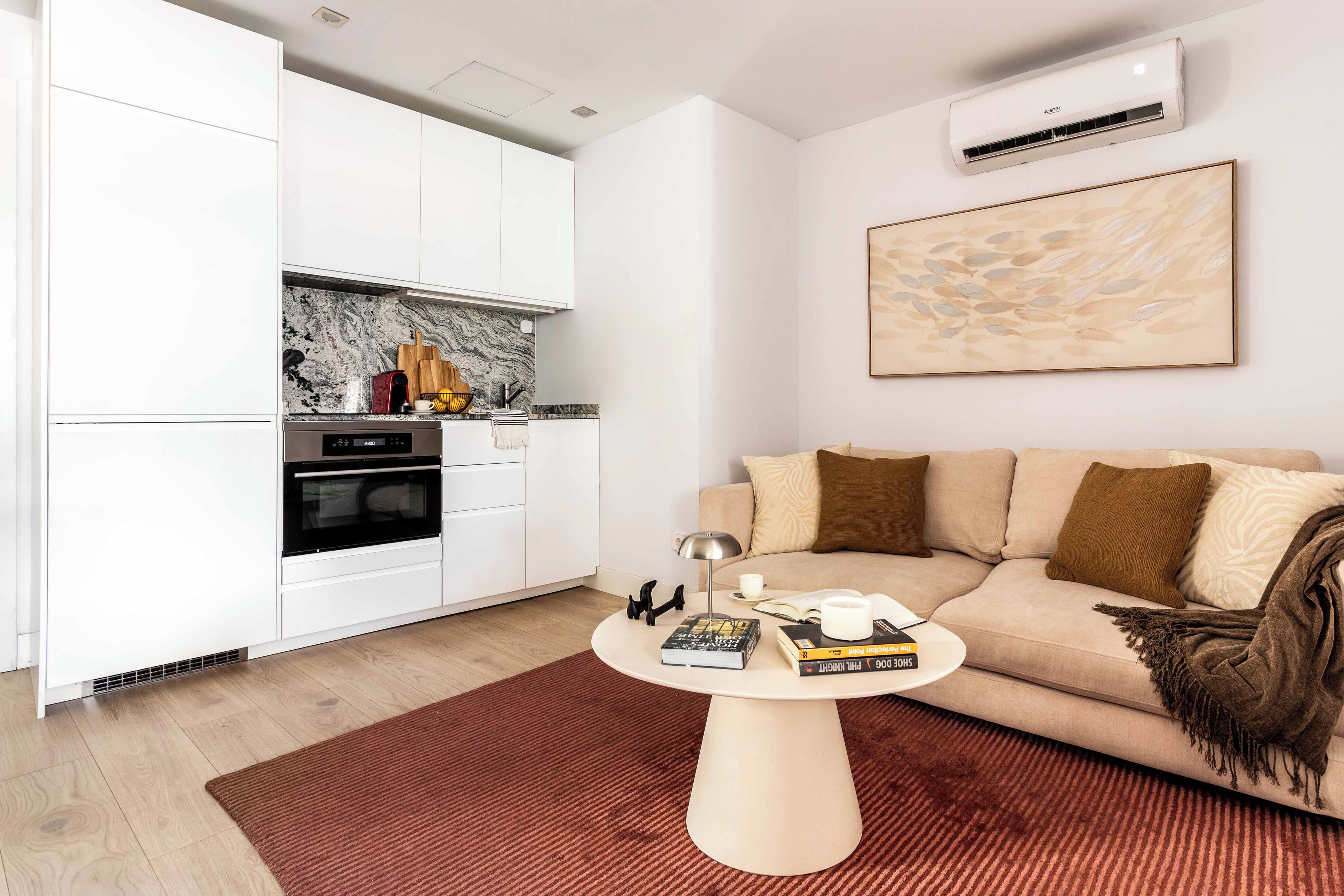 Una sala de estar moderna con un sofá color beige, una mesa de centro y una zona de cocina con armarios blancos y salpicadero de mármol.