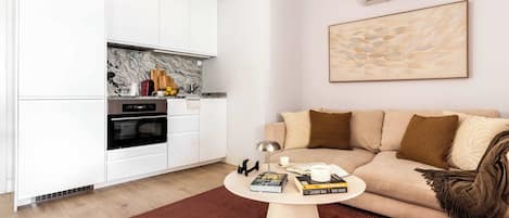 Una sala de estar moderna con un sofá color beige, una mesa de centro y una zona de cocina con armarios blancos y salpicadero de mármol.