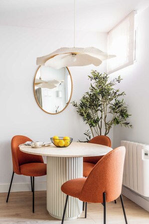 A dining area with a round table, two chairs, a bowl of fruit, a potted plant, and a wall-mounted mirror. - This elegant one-bedroom apartment in the heart of Justicia—one of Madrid’s most vibrant and exclusive neighborhoods—combines contemporary design, comfort, and a sophisticated atmosphere ideal for extended stays (MADRID)