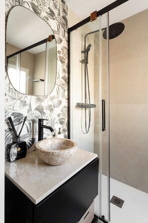 A bathroom with a marble sink, a black faucet, a glass shower enclosure, and a mirror with a floral wallpaper. - This elegant one-bedroom apartment in the heart of Justicia—one of Madrid’s most vibrant and exclusive neighborhoods—combines contemporary design, comfort, and a sophisticated atmosphere ideal for extended stays (MADRID)