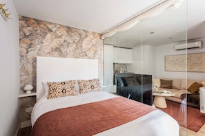 A modern hotel room with a large bed, a glass partition, a sofa, and an air conditioning unit. - This elegant one-bedroom apartment in the heart of Justicia—one of Madrid’s most vibrant and exclusive neighborhoods—combines contemporary design, comfort, and a sophisticated atmosphere ideal for extended stays (MADRID)