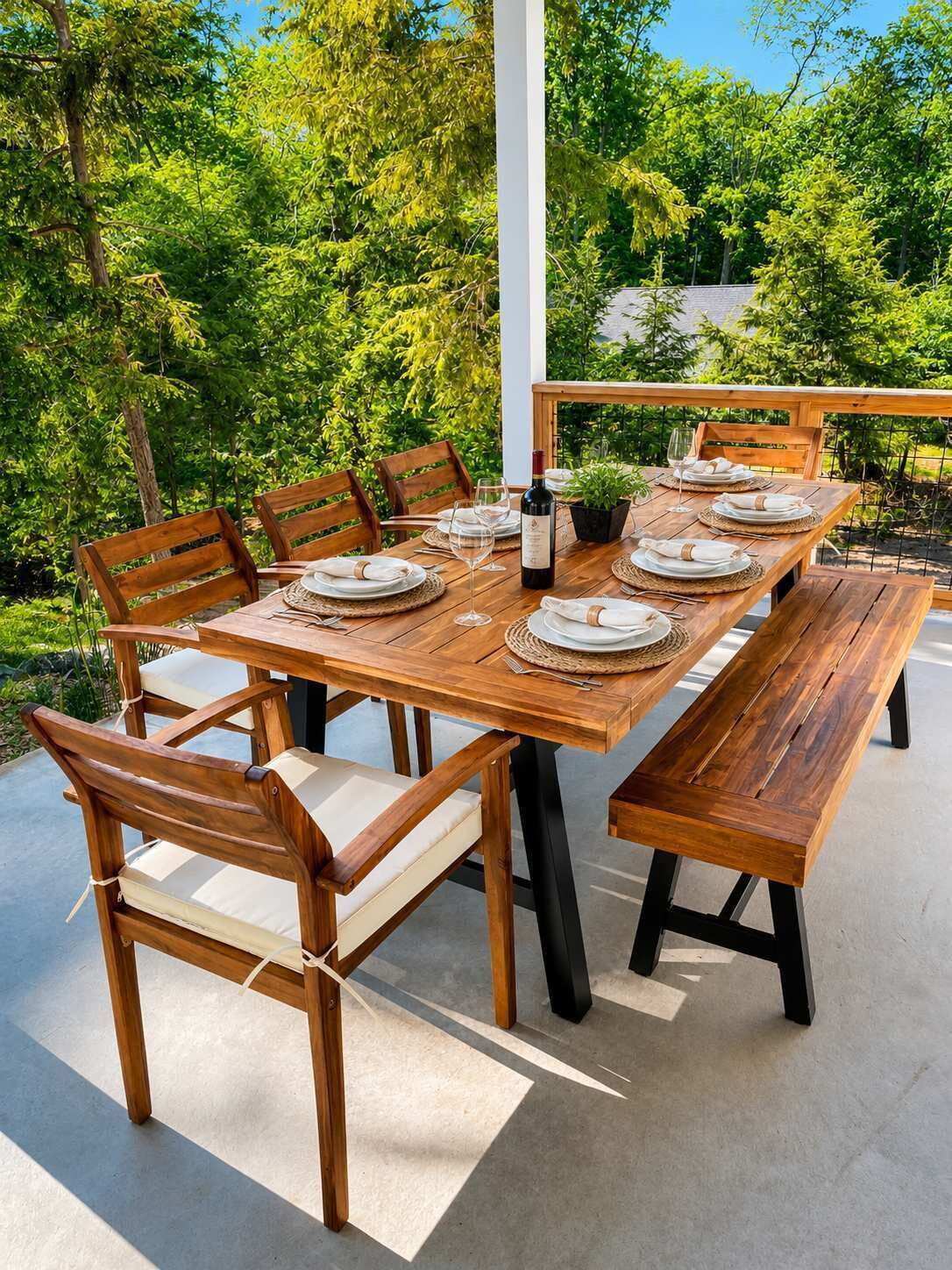 An outdoor wooden dining table set with chairs, plates, and a bottle of wine, surrounded by lush greenery.