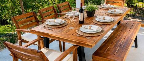 An outdoor wooden dining table set with chairs, plates, and a bottle of wine, surrounded by lush greenery.