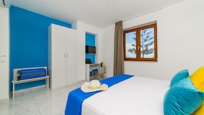 Ett hotellrum med en säng, ett skrivbord, en garderob, en bänk och ett fönster med utsikt över träd.