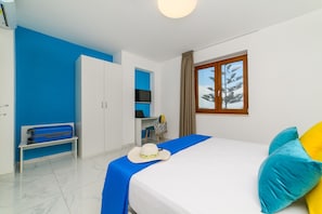 A hotel room with a bed, a desk, a wardrobe, a bench, and a window with a view of trees. - Iris Flat (Massa Lubrense)