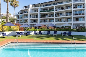 Una piscina con sillones de descanso y sombrillas, rodeada de un césped cuidado y un edificio de varios pisos con balcones.