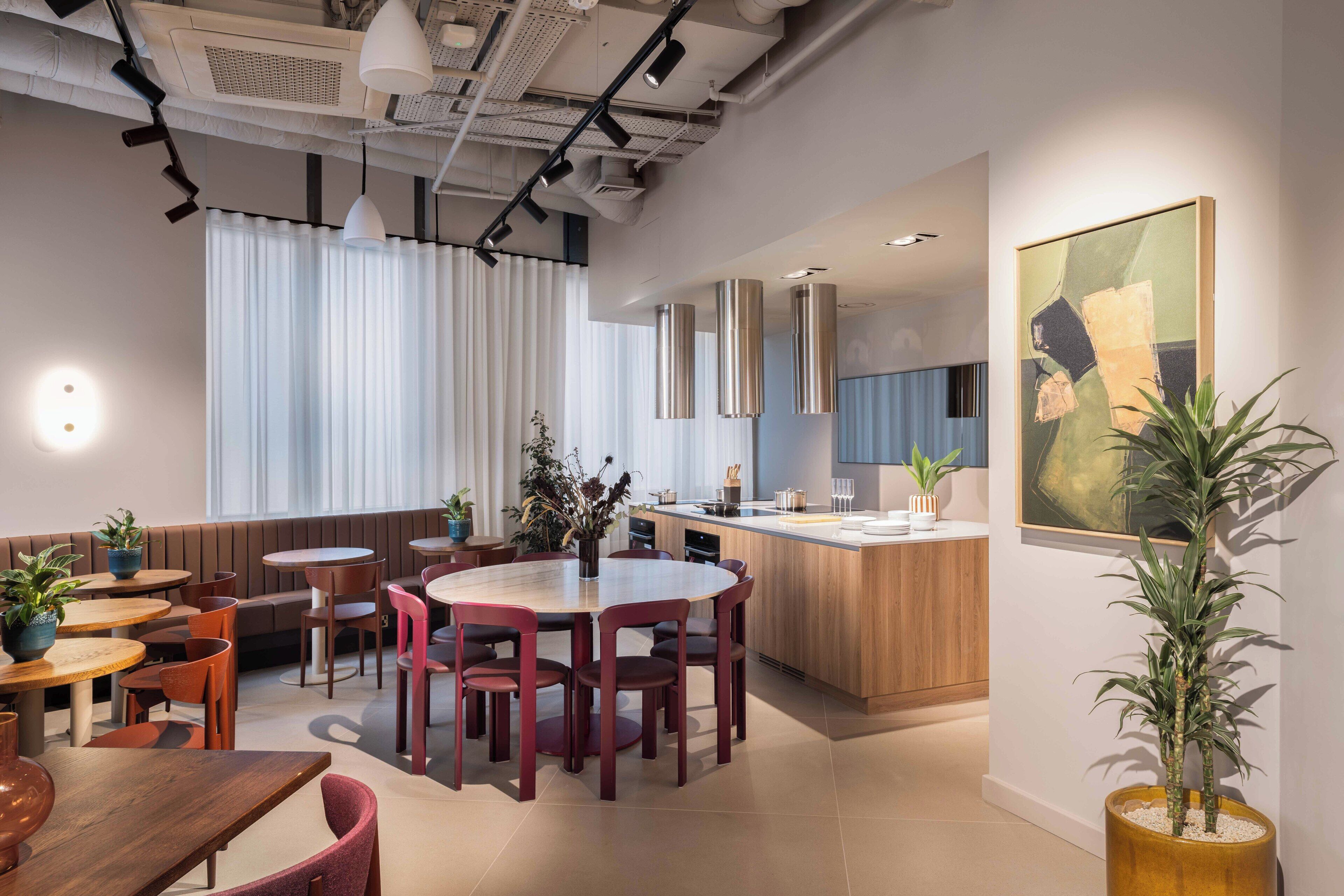 A modern dining area with round tables and chairs, a kitchen area, and a large painting on the wall.