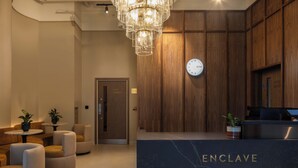 A reception area with a marble counter labeled 'ENCLAVE,' wooden panel walls, a chandelier, and seating with potted plants. - StayCozy Studio Apartments in Acton (London)
