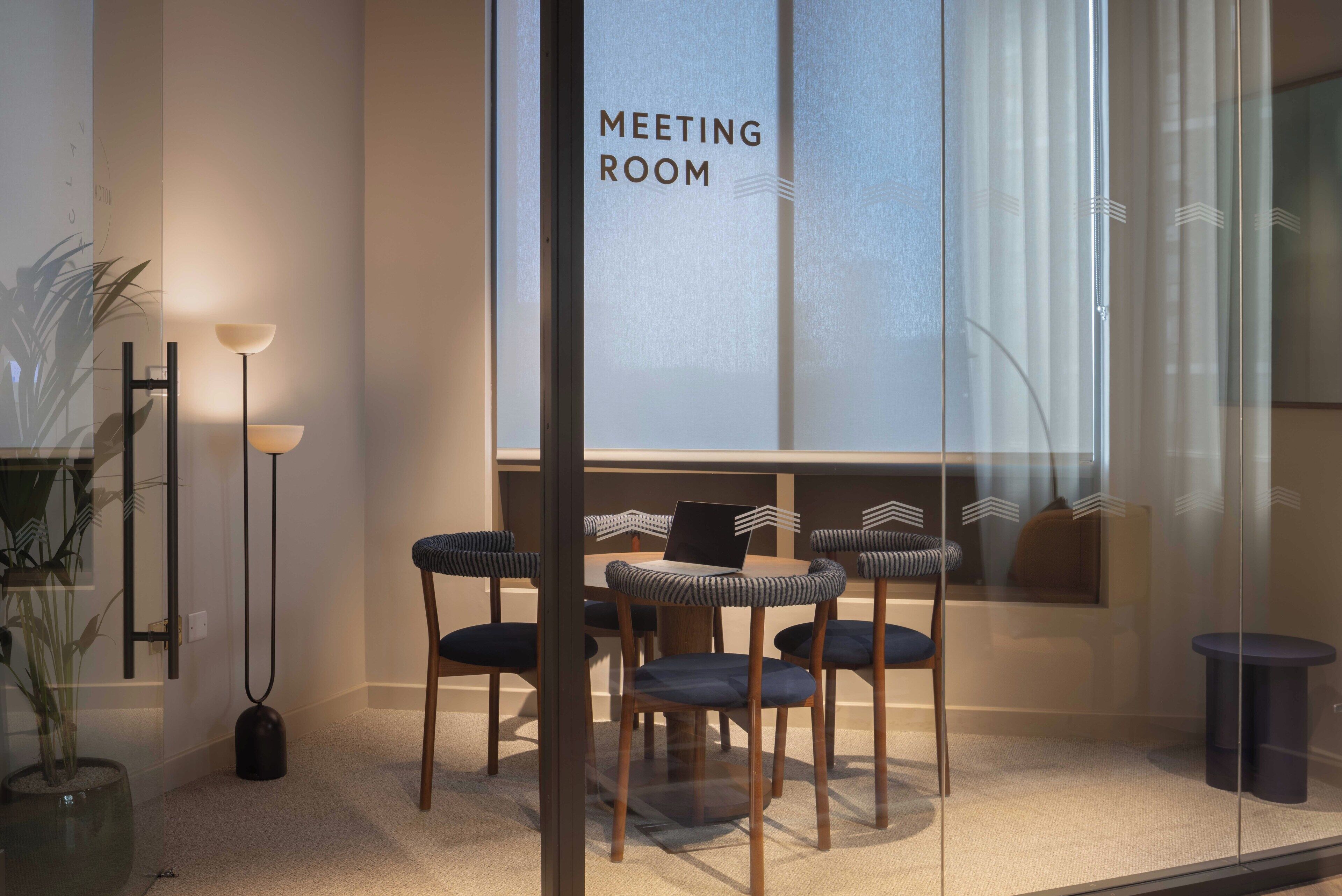 A meeting room with a glass partition, a table with chairs, a floor lamp, and a potted plant.
