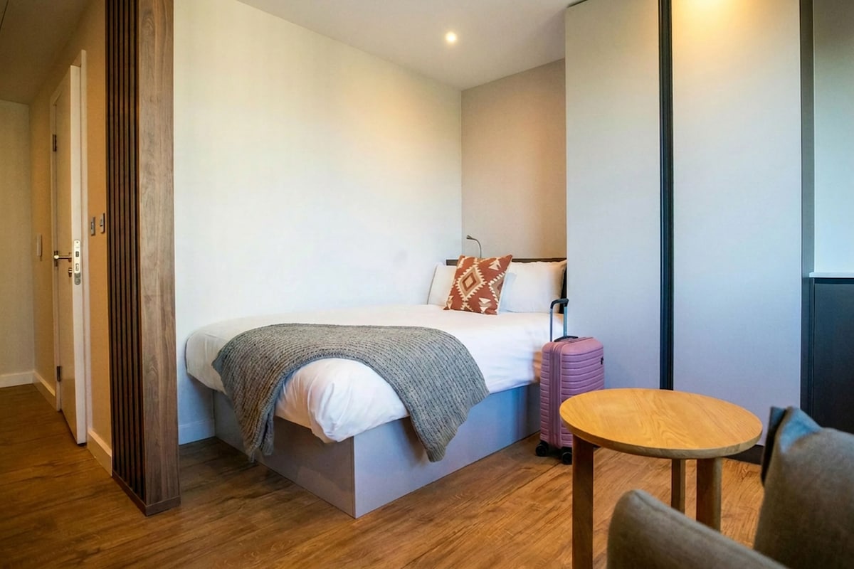 A bedroom with a bed, a bedside table, a suitcase, and a door.