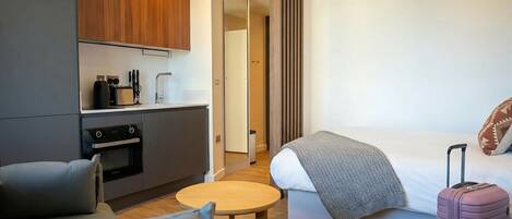 A modern hotel room with a kitchenette, a bed with a blanket, a grey sofa, and a suitcase.
