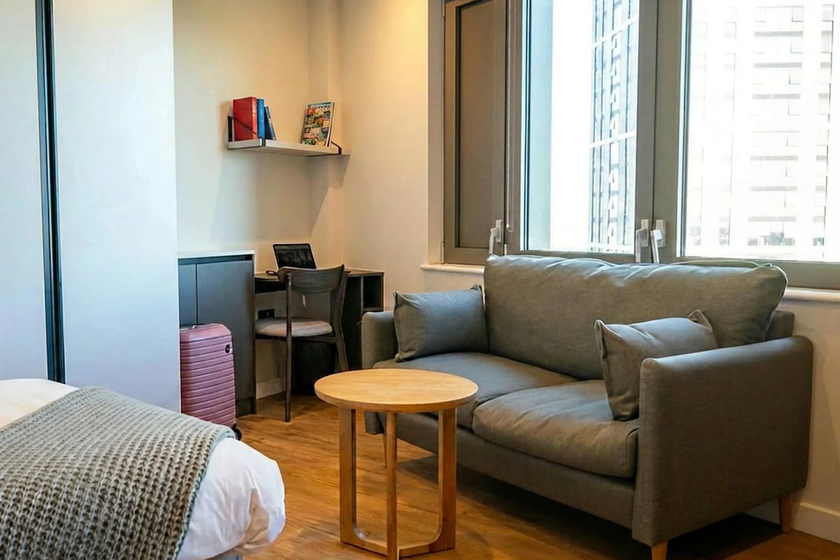 A modern hotel room with a sofa, a small round table, a desk with a laptop, a pink suitcase, and a bookshelf with books and a picture frame.