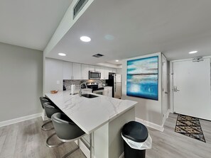 A modern kitchen with white cabinetry, a marble countertop, and a black trash bin. - Edgewater #1302 | Gulf Front + Amenities (Miramar Beach)