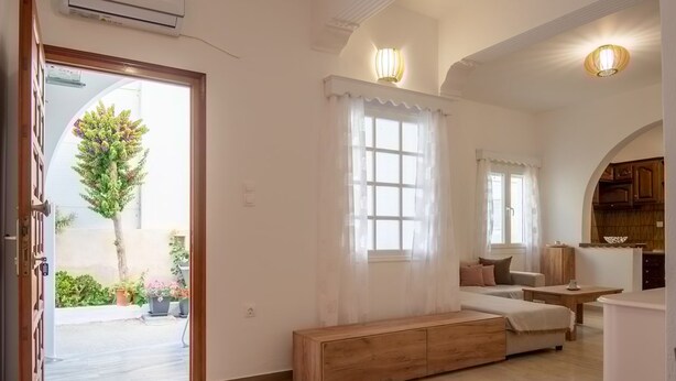 A spacious living room with a wooden bench, a sofa, and a dining area. There is a large window with curtains, and a view of a tree and plants outside. - Naousa Central House, Paros (Paros)