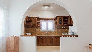 A kitchen with wooden cabinets, a window with a lace curtain, and a countertop with a kettle and a phone. - Naousa Central House, Paros (Paros)