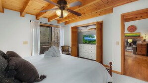 Ein Schlafzimmer mit Bett, Deckenventilator, einem Fenster mit Vorhängen, einer Holztür, die ins Badezimmer führt, und Blick auf eine Landschaft durch eine Glastür.
